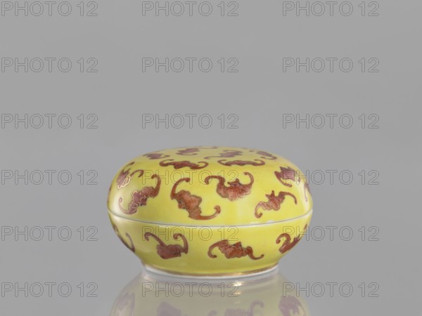 Round box with a convex lid a cinnabar vessel for seals, bats iconography, ceramics, colored porcelain, Qing style, Tongzhi 1862–1874, Chinese, lids, seals, Qing 1644–1911, 1862–1874, China, vessel, height 4 cm, diameter 7 cm, A small round box with a lid sits centered against a plain gray background, the box painted a bright yellow color and decorated all over with a repeating organic motif in a reddish-brown color; the box has a smooth glossy surface and a thin pale rim where lid and base meet, and its reflection is faintly visible on the surface beneath it.