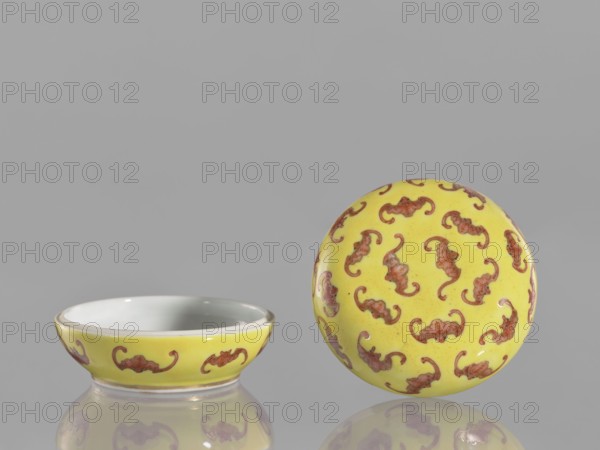 Round box with a convex lid a cinnabar vessel for seals, bats iconographic, ceramics, colored porcelain, Qing style, Tongzhi 1862–1874, Chinese culture, lid, seals, Qing 1644–1911, 1862–1874, China, vessel, height 4 cm, diameter 7 cm, A shallow open bowl and a matching lid sit on a reflective surface against a gray background; both pieces are primarily bright yellow with a repeating decorative motif in reddish-brown, the bowl’s interior is glossy white, and the lid shows the same yellow and reddish-brown pattern across its rounded top, with reflections of the objects visible on the surface below.