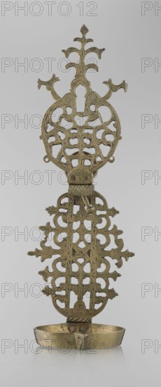 Oil lamp, Persian/Islamic art, 19th–20th century, Central Asia or Iran, oil lamp with side decorative shaft, stylized medallion and palmette finial; decorated with birds, geometric motifs and palmettes; height 45.5 cm, diameter 15.5 cm., A vertical metal object with an ornate openwork design and a shallow bowl at the base, shown against a plain light gray background; the metal has a muted gold to brass color with areas of darker patina and slight discoloration; the top portion features symmetrical curving branches and rounded cutouts forming a decorative crest, followed by a central section with repeating open scrollwork and stylized floral or foliate motifs; a small hinge-like element connects the upper and lower panels, and a few small protruding pins or rivets are visible where the bowl meets the main structure; the shallow bowl at the bottom is oval with a gently curved lip and shows subtle surface marks and a slightly uneven sheen.