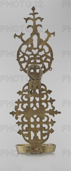 Oil lamp, Persian, 19th–20th century, possibly Central Asia or Iran; ceramic/metal oil lamp with side decorative shaft, stylized medallion and palmette finial; decorated with birds, geometric motifs, and palmette ornament; height 45.5 cm, diameter 15.5 cm., An upright metal object with an ornate openwork design and a hinged ring near the top, featuring a decorative top section with branching shapes and a larger symmetrical middle section of repeating cutout patterns, all showing a mottled metallic gold and brown patina; a small rectangular hinge piece connects to a lower ornamental panel with similar cutout motifs, which sits above a shallow metal tray with a slightly curved rim that shares the same gold and brown coloring; the background is light gray.