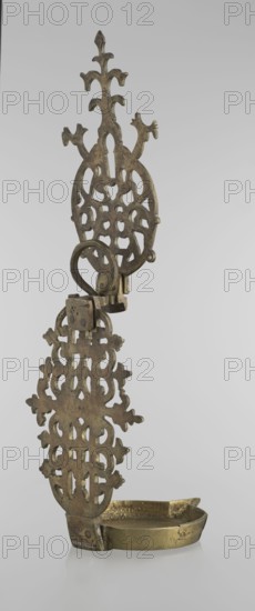 Oil lamp, gift, Islamic art, Persian, oil lamp for lighting decorated with birds, geometric motifs, palmettes; lamp with a side decorative shaft, stylized medallion, palmette finial. 19th–20th century, Central Asia, / Iran, height 45.5 cm, diameter 15.5 cm, An ornate metal oil lamp with a vertical, pierced decorative backplate featuring an intricate, symmetrical openwork pattern and fleur-like extensions, attached to a hinged ring and bracket above a shallow oval basin; the object shows a metallic surface in shades of brass and dark brown with areas of patina and slight wear against a pale gray background.