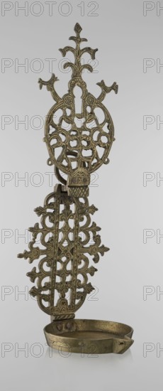 Oil lamp, gift, Islamic art, Persian culture, oil lamp for lighting decorated with birds, geometric motifs, palmettes; lamp with a side decorative shaft, stylized medallion and palmette finial. 19th–20th century, Central Asia, Iran, height 45.5 cm, diameter 15.5 cm., An ornate metal oil lamp with an elongated decorative backplate featuring a lattice of interlocking scrollwork and floral motifs in a brass or bronze tone, the backplate rising into a stylized treelike finial with curved branches, and a shallow rounded reservoir at the base with a small spout, all shown against a plain light gray background.