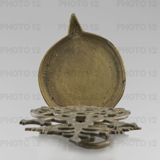 Oil lamp, Persian/Islamic art, oil lamp for lighting with birds, geometric motifs, palmettes; lamp with a side decorative shaft, stylized medallion and palmette finial. 19th–20th century, Central Asia, / Iran, height 45.5 cm, diameter 15.5 cm., A round, shallow metal dish with a pointed protrusion at the top is shown upright, revealing a textured, worn interior surface in a brass-like golden color; in front of it lies an ornate flat metal piece with openwork patterns and rounded protrusions, also in a similar golden-brass color with darker patina and worn edges, all set against a smooth light gray background with reflections visible beneath the objects.