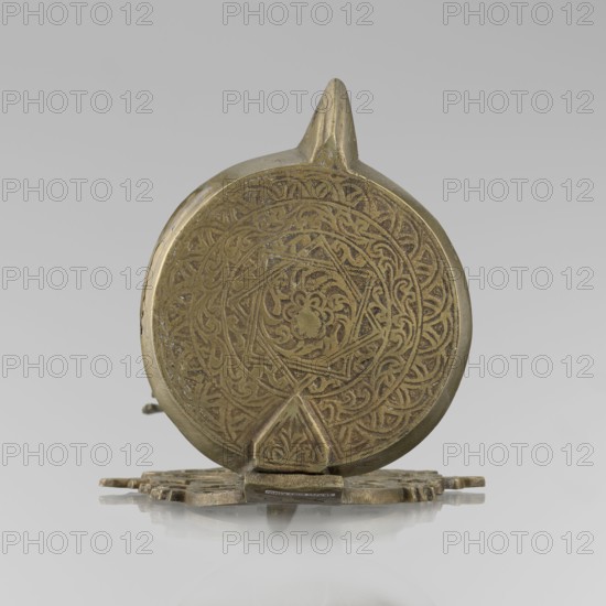 Oil lamp, Islamic art, Persian culture; oil lamp with lighting function decorated with birds, geometric motifs, palmettes ornament; lamp with a side decorative shaft, stylized medallion, palmette finial. 19th–20th century, possibly Central Asia or Iran. Height 45.5 cm; diameter 15.5 cm., A circular metallic object with an embossed ornate pattern on its face sits upright on a flat reflective surface; the object appears in shades of gold and brass with darker engraved lines creating a swirling floral and geometric design, and a small triangular projection at the bottom and a pointed fin at the top.