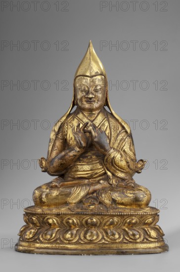 Gelukpa teacher, unknown sculptor, 18th century, Tibet; Tibetan Buddhist Himalayan sculpture; mudras; lotus throne iconography; headgear; height 22.5 cm, width 16.5 cm., A gilded seated figure wearing a pointed headdress and layered robes sits on a rounded lotus base; the figure's hands are held together at chest level in a precise gesture, the facial features are calm with closed eyes and a slight smile, and a few darker worn areas interrupt the gold finish. The sculpture shows a mix of gold and darker bronze tones on the body and base, with subtle shadows on the drapery folds and ornamental patterns on the sleeves and base. The background is a flat neutral gray.