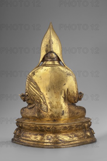 Gelukpa teacher, unknown sculptor, 18th century, Tibet; Tibetan Buddhist Himalayan sculpture; mudras; lotus throne iconography; headgear; height 22.5 cm, width 16.5 cm., A gilded statue shown from the back on a round ornamental base, with a pointed hooded headpiece and draped shoulders; the surface shows areas of worn gold with darker spots and patina, and a decorative patterned cloth hanging from one shoulder; the base features carved leaf and bead motifs. Colors visible include gold, darker brownish tarnish, and a neutral gray background.
