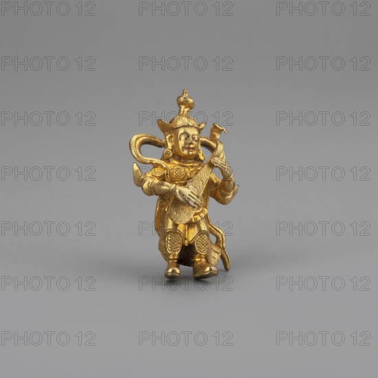 Dhritarashtra, Himalayan culture, Tibetan Buddhist art, Himalayan art, musical instrument, 2nd half of the 18th–19th century, Mongolia, Tibet, sculpture, figurine, cult object, height 3.4 cm, A small golden figurine of a person seated with legs bent, wearing an ornate headdress and layered clothing, holding and plucking a stringed instrument; the figure has rounded facial features and detailed decorative elements on the clothing and armor, with curved ornamental extensions behind the head and a small rounded finial atop the headdress. The scene shows a plain neutral gray background and gray surface beneath the figurine; the visible colors are gold on the figurine and shades of gray in the background and surface.