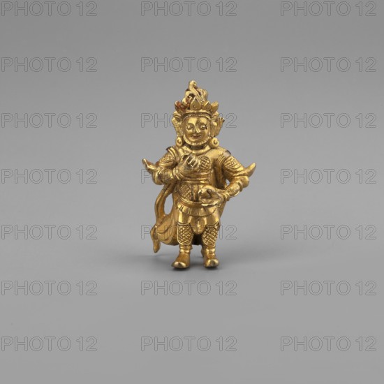 Vaisravana Tib. Namtose, Tib. rnam thos sras, sometimes identified with the yellow Jambhala, an emanation of Buddha Ratnasambhava — guardian of the north. Himalayan Tibetan Buddhist art. 2nd half of the 18th–19th century. Possibly Mongolia or Tibet. Sculpture; figurine; cult object. Height: 3.4 cm., A small gold-colored figurine stands on a gray surface against a neutral gray background, depicting a robed figure wearing ornate headgear and layered clothing with textured patterns; the figure has a rounded face with visible facial features, articulated arms with one hand near the chest and the other extended forward holding a small object, decorative elements around the shoulders and waist, a flowing sash or drape at the back, and detailed leg coverings with patterned design.