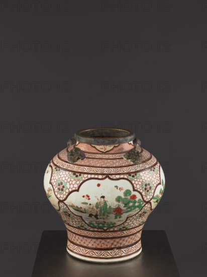 Vase, Chinese, Qing 1644–1911, mid-18th century. Colored porcelain vessel, height 40.5 cm, diameter 40 cm. Mascarons on the shoulder; decoration: four reserves with figural representations and gilded edge., A rounded ceramic jar sits on a dark surface against a dark gray background, decorated with intricate painted patterns and figurative scenes; the jar has a wide neck with a darker rim and small sculpted lion-head handles near the top. The body displays scalloped panels outlined in reddish-brown that contain painted human figures and stylized trees and foliage, with a palette that includes white, reddish-brown, green, and touches of black and pale blue. Repeating geometric and wave-like bands in reddish-brown encircle the neck and base, and floral or lattice motifs fill spaces between the panels. The overall visual impression emphasizes ornate, symmetrical decoration with contrasting areas of white and colored ornamentation.