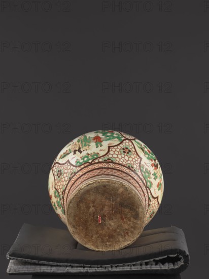 Vase, Chinese, mid-18th century Qing dynasty, 1644–1911. Colored porcelain vessel, height 40.5 cm, diameter 40 cm. Mascarons on the shoulder; decoration: four reserves with figural representations; gilded edge., A round ceramic vessel resting on folded dark gray and black cushions, shown from a low angle with its base facing the viewer; the vessel has an off-white ground with painted decorative bands and panels in green, red, and brown, including floral and geometric patterns and small painted human figures, and the exposed base appears worn and brownish with a small red mark.