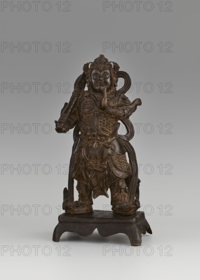 Weituo — protector of Buddhist learning and monasteries., Figurine, sculpture, 15th/16th century, China. Materials/technique: sculpture. Height 32.0 cm, width 15.8 cm, depth 10.5 cm. Subject/iconography: Weituo a bodhisattva and guardian deity in Chinese Buddhism depicted as a general wearing a high helmet and full chain mail armor, belt adorned with a monster's head, usually shown with a sword; traditionally said to have fought demons that tried to steal Buddha's relics. Categories: Chinese art; Chinese Buddhism; guardian deities; generals; figurines., A dark brown carved figure stands on a low pedestal, wearing detailed armor and layered garments with elaborate patterns and textured surfaces. The figure holds a long cylindrical object in one hand while the other hand is raised near the face, and flowing ribbon-like elements wrap around the torso and extend beside the legs. The face shows prominent facial features and a headdress with ornamental details. The pedestal is also dark brow...