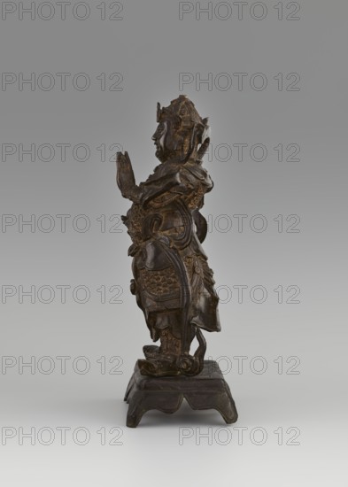 Weituo — protector of Buddhist learning and monasteries; unknown creator; figurine; Chinese art; Chinese Buddhism; guardian deity/bodhisattva iconography. Weituo preserves Buddhist teachings and protects monasteries; traditionally fought demons that tried to steal Buddha's relics, hence depicted as a general wearing a high helmet and full chain mail armor, belt adorned with a monster's head, usually shown with a sword. 15th–16th century, China. Sculpture; height 32.0 cm, width 15.8 cm, depth 10.5 cm., A dark brown carved figure stands on a carved base against a neutral gray background, shown in profile with hands held together near chest level. The figure wears an ornate headdress and layered armor with textured detailing, including scale-like patterns and flowing drapery elements that extend down the sides. The surface shows areas of lighter brown highlights on raised details. The base is a darker brown with a slightly weathered appearance and curved cutouts along its lower edge. The ...