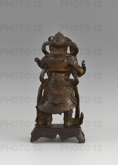 Weituo — protector of Buddhist learning and monasteries;, figurine; Chinese art; Chinese Buddhism; guardian deity and bodhisattva iconography. According to tradition, Weituo's role is to preserve Buddhist teachings and protect monasteries; he fought demons attempting to steal Buddha's relics and is depicted as a general wearing a high helmet and full chain mail armor, secured at the waist with a belt adorned with a monster's head, usually shown with a sword. Sculpture, 15th–16th century, China. Dimensions: height 32.0 cm; width 15.8 cm; depth 10.5 cm., A dark brown carved figure viewed from behind, standing on a low scalloped base; the figure wears layered armor with textured scale-like patterns and draped skirt elements, and has looping ornamental bands or sashes that curve around the shoulders and back; the head is topped with a rounded, decorated helmet or headdress, and the arms are bent with hands extending slightly outward; the surface shows polished highlights and worn areas in ...