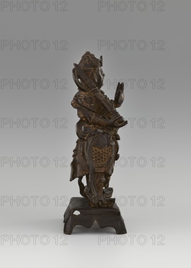 Weituo — protector of Buddhist learning and monasteries., Figurine, sculpture, 15th/16th century, China. Chinese art; Chinese Buddhism; guardian deity/bodhisattva iconography; generals. Weituo is a protector whose task is to preserve Buddhist teachings and protect monasteries; according to tradition he fought demons that tried to steal Buddha's relics and is depicted as a general wearing a high helmet and full chain mail armor, secured at the waist with a belt adorned with a monster's head, usually shown with a sword. Dimensions: height 32.0 cm; width 15.8 cm; depth 10.5 cm., A carved standing figure on a small carved pedestal, shown in profile, with ornate armor and layered garments, a headdress, and hands raised in a gesture; the carving has visible carved patterns and flowing drapery elements, and the surface appears dark brown with some lighter brown highlights, all set against a neutral gray background.