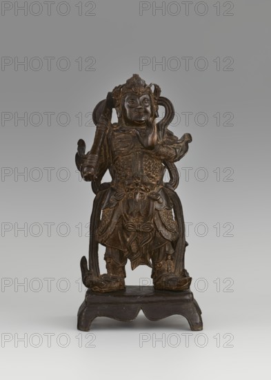 Weituo — protector of Buddhist learning and monasteries., Figurine, Chinese, Chinese Buddhism. Guardian deity/bodhisattva iconography: Weituo preserves Buddhist teachings and protects monasteries; tradition says he fought demons trying to steal Buddha's relics and is depicted as a general wearing a high helmet and full chain-mail armor, secured at the waist with a belt adorned with a monster's head, usually shown with a sword. Sculpture, 15th–16th century, China. Dimensions: height 32.0 cm; width 15.8 cm; depth 10.5 cm., A carved statue of a standing figure is centered on a raised base against a neutral background; the figure appears in dark brown tones with lighter brown highlights on carved details, and there are traces of golden or gilt color in some of the raised areas. The figure wears layered, ornate armor and draped decorative elements that cascade down to the feet, with sculpted patterns and relief work across the torso and skirt area. One hand is raised near the face while the...