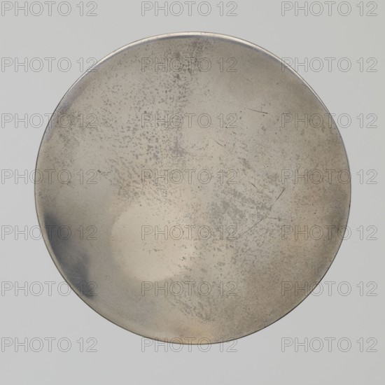 Mirror, Tang Dynasty 8th century, 618–907, China. Bronze mirror with beasts fantastical, frogs, birds, and vine motifs. Diameter 9.8 cm; height 0.9 cm., A circular metallic object fills the frame, showing a worn, mottled surface with patches of light beige, pale grey, silvery tones, and areas of darker grey; the rim appears slightly darker and metallic, and there are subtle scratches and speckled spots across the surface creating a textured, uneven appearance.