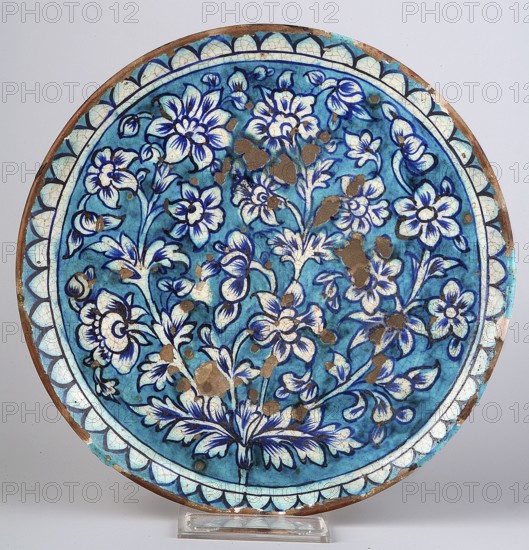 Plate;, Ceramic vessel with height 3 cm and diameter 26.5 cm. 19th century, India/Pakistan . Decoration: composition of a stylized flower bush and crenellated ornament at the edge; floral motifs; plant ornament. Islamic/Indian art., A round ceramic plate with a blue background displaying a floral pattern in darker blue and white tones, with outlines and details in a deep blue or black. The central area shows a cluster of stylized flowers and leaves radiating outward, with petals and foliage rendered in white and blue shading. The rim features a decorative border of scallop-like shapes in pale blue and white with dark blue outlines, and there are several areas of brown spots and worn patches across the surface revealing the underlying material. The plate rests on a clear display stand against a light neutral background.