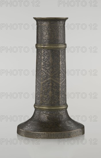 Candlestick mash'al, Islamic, Persian, Safavid dynasty 1501–1736; 17th–18th century, Iran. Metal candlestick in the form of a column standing on a low, widening base with three sections separated by hachured rings. Decoration: 10-faceted shaft with stylized plant ornament and a zigzag; band of nast'alik inscriptions just under the upper edge; hunting scenes beneath, background filled with a dense tendril pattern; base decorated with huntsmen and an edge inscription in Armenian characters of decorative value. Height 28.0 cm; diameter 17.3 cm; weight 0.71 kg., A tall cylindrical metal object with a flared base stands against a light gray background. The surface shows intricate engraved and etched ornamentation with repeating floral and geometric patterns and some circular medallion designs. The object has a darker brownish-gray patina with areas of muted golden-bronze highlighting on raised bands and edges. There are a few horizontal ridged bands of brighter golden-bronze encircling the ...
