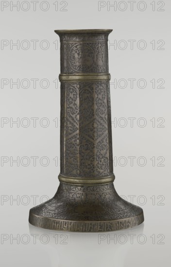 Candlestick mash'al, Islamic art, Persian, Safavid dynasty 1501–1736. 17th/18th century, Iran. Bronze candlestick in the form of a column standing on a low, widening base with three sections separated by hachured rings. Ten-faceted shaft decorated with a stylized plant ornament and zigzag. Just under the upper edge is a band of nast'alik script; beneath this are hunting scenes and a dense tendril background. The base is decorated with figures of riders/huntsmen; an Armenian-script inscription on the base edge appears decorative. Height 28.0 cm; diameter 17.3 cm; weight 0.71 kg., The image shows a tall, tapered metal object with a flared base, decorated all over with intricate engraved patterns and repeated decorative bands; the surface appears dark bronze with areas of lighter golden highlights, and there are a few raised horizontal ridges with a greener gold tone encircling the form; the engravings include ornamental vine-like motifs and several medallion-shaped panels, and the overal...
