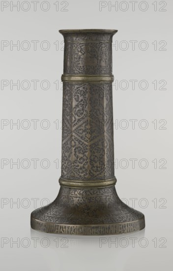 Candlestick mash'al, Islamic art, Persian, Safavid dynasty 1501–1736; 17th–18th century, Iran. Bronze candlestick in the form of a column standing on a low widening base, three sections separated by hachured rings. Ten-faceted shaft decorated with stylized plant ornament and a zigzag. Just under the upper edge is a band of inscriptions in nast'aliq script; beneath this are hunting scenes with riders and horses set against a dense tendril pattern. The base is decorated with representations of huntsmen; an inscription in Armenian characters runs on the edge and serves a decorative function. Height 28.0 cm; diameter 17.3 cm; weight 0.71 kg., A tall cylindrical metal object with a flared base and a narrower top, displaying intricate engraved floral and geometric patterns across its surface; the metal appears dark with areas of golden or brass-colored highlights on raised bands and decorative edges, and a few horizontal ridged bands encircle the cylinder; the surface shows repeated vine-lik...