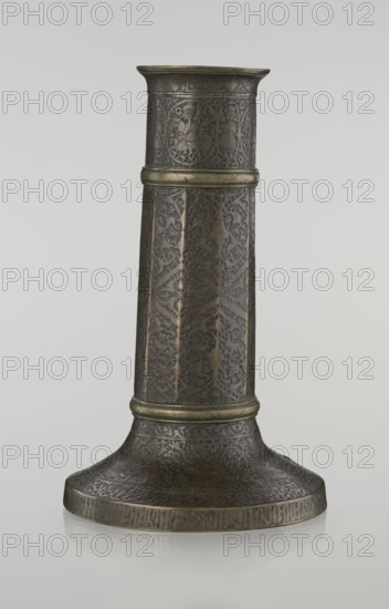 Candlestick mash'al, Islamic art, Persian, Safavid dynasty 1501–1736. Metal candlestick in the form of a column standing upon a low, widening base, its three sections separated by hachured rings. The 10-faceted shaft is decorated with stylized plant ornament and a zigzag; just under the upper edge is a band of inscriptions in nast?aliq script; beneath this are hunting scenes with riders and horses; the background filled with a dense tendril pattern. The base is decorated with representations of huntsmen; an inscription in Armenian characters appears on the base edge, serving a decorative function. 17th–18th century, Iran. Height 28.0 cm; diameter 17.3 cm; weight 0.71 kg., An upright cylindrical metal object with a flared base, shown against a plain light gray background; the object is predominantly dark bronze with areas of muted gold detailing, and its surface is covered in intricate carved or etched repeating patterns and decorative motifs. The midsection and upper rim have raised ba...