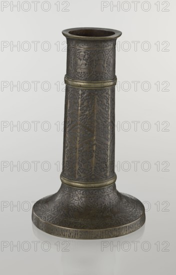 Candlestick mash'al, Islamic, Persian, Safavid dynasty 1501–1736. Candlestick in the form of a column standing upon a low, widening base, its three sections separated by hachured rings. Decoration: stylized plant ornament and a zigzag on the 10-faceted shaft; band of nast'aliq inscriptions just under the upper edge; hunting scenes beneath; background filled with a dense tendril pattern. Base decorated with representations of huntsmen; an inscription in Armenian characters on the base edge of decorative value., metalware, 17th–18th century, Iran. Dimensions: height 28.0 cm; diameter 17.3 cm; weight 0.71 kg., A tall cylindrical metal object with a flared round base sits against a plain light gray background; the surface shows intricate embossed and engraved decorative patterns covering most of the piece, with a few horizontal raised bands encircling the cylinder that are a slightly brighter yellowish metal; the overall color palette includes dark brown and bronze tones with areas of mute...