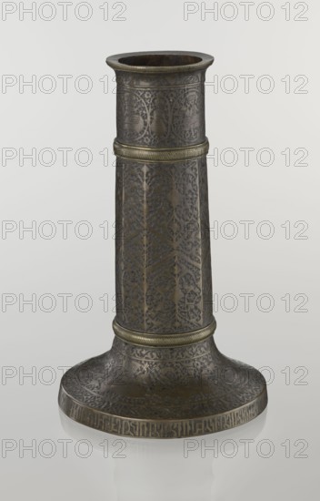 Candlestick mash'al, Islamic art, Persian, Safavid dynasty 1501–1736. Metal candlestick in the form of a column standing upon a low, widening base, its three sections separated by hachured rings. The 10-faceted shaft is decorated with a stylized plant ornament and a zigzag. Just under the upper edge is a band of inscriptions in nast'aliq script; beneath this are hunting scenes with riders and horses set against a dense tendril pattern. The base is decorated with representations of huntsmen; an inscription in Armenian characters appears on the base edge and serves a decorative function. 17th–18th century, Iran. Height 28.0 cm; diameter 17.3 cm; weight 0.71 kg., A tall, cylindrical metal object with a flared base, shown against a plain light gray background; the surface is dark bronze with areas of lighter antique brass highlights, and it is decorated all over with intricate engraved floral and geometric patterns and a few bands of raised ridged trim in a brass tone; the top rim is sligh...