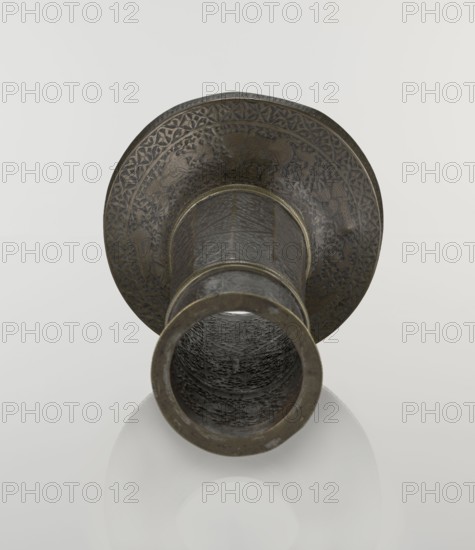 Candlestick mash'al, Islamic art, Persian, Safavid dynasty 1501–1736, 17th–18th century, Iran. Bronze candlestick in the form of a column standing upon a low widening base, its three sections separated by hachured rings. Decoration: 10-faceted shaft with stylized plant ornament and a zigzag; band of inscriptions in nast'alik script just under the upper edge; hunting scenes beneath, the background filled with a dense tendril pattern; base decorated with representations of huntsmen and figures of riders on horses in the upper part. Stylized inscriptions in Persian and an Armenian inscription on the base decorative. Function: candlestick. Height 28.0 cm; diameter 17.3 cm; weight 0.71 kg., An ornate metal object viewed from one open end, showing a cylindrical interior with a textured dark surface and a wider flared rim decorated with intricate engraved patterns; the metal appears in shades of dark brown and bronze with areas of darker gray, and the background is a smooth off-white.