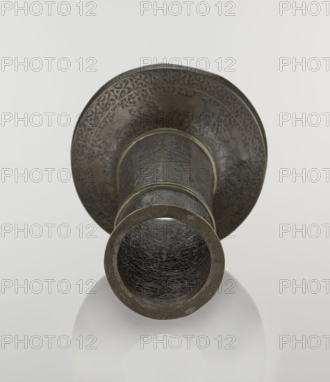 Candlestick mash'al, Islamic art, Persian, Safavid dynasty 1501–1736. Candlestick in the form of a column standing upon a low, widening base; three sections separated by hachured rings. Just under the upper edge, a band of inscriptions in nast'alik script; beneath this, hunting scenes. Background filled with a dense tendril pattern. Base decorated with representations of huntsmen; upon its edge an inscription in Armenian characters of decorative value. Decoration: stylized plant ornament and a zigzag on the 10-faceted shaft, figures of riders at the base and in the upper part, stylized inscriptions: Persian and Armenian. 17th/18th century, Iran. Candlestick; height 28.0 cm, diameter 17.3 cm, weight 0.71 kg., A metallic object viewed from the front showing a flared rim and a cylindrical body, with a textured interior visible through the opening. The surface displays engraved or embossed decorative patterns, and there are visible ridged bands around the cylinder. The object appears in sh...