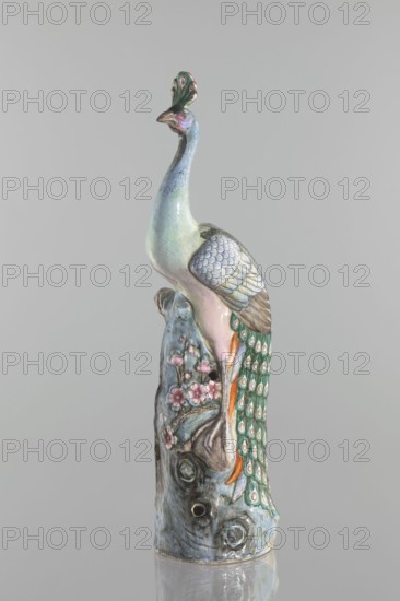 Peacock figurine, Chinese, Qing dynasty 1644–1911, 19th century. Ceramic colored porcelain sculpture depicting a peacock with peacock feather ornament, images of birds, plum tree and tree trunk with flowers. Height 54.5 cm, width 14.0 cm, depth 12.5 cm, weight 2.9 kg., A ceramic sculpture of a bird perched on a textured base, showing a long neck and an upright crest; the bird displays a pastel palette with pale blue and green tones on its body, pinkish hues on its chest and face, and brown shading on its beak and wing tips. The wing features overlapping scale-like feathers in muted blue and gray, and a long tail cascades down the base with green and teal feathers highlighted by circular eye-like patterns in cream and gold and small accents of orange. The base resembles weathered wood or rock with blue-gray and brown tones and is decorated with a few pink blossoms and branches, and the entire piece sits against a neutral light gray background with a subtle reflective surface beneath it.