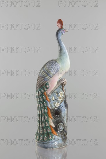 Peacock figurine, Chinese, Qing dynasty 1644–1911, 19th century. Ceramic colored porcelain sculpture depicting a peacock with peacock-feather ornament, images of birds, plum trees, tree trunks, and flowers. Height 54.5 cm; width 14.0 cm; depth 12.5 cm; weight 2.9 kg., A ceramic sculpture of a bird perched on a textured base, shown against a plain pale gray background. The bird has a long curved neck and a small head topped with a crest that is reddish-orange. Its body shows soft gradients of pale green, bluish-gray, and light pink, with layered wing feathers rendered in muted blue-gray tones. A flowing tail cascades down the base with eye-like feather patterns in green, teal, and cream, accented with orange feathers. The base has a marbled and weathered appearance in blue-gray and darker gray, with small raised floral decorations in cream and pale yellow and a few circular openings. The overall color palette includes pale green, blue-gray, teal, green, orange, cream, pale yellow, pink,...