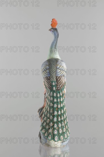 Peacock figurine, Chinese, Qing dynasty 1644–1911, 19th century. Colored porcelain sculpture depicting a peacock with peacock-feather ornament, images of birds, plum trees, tree trunks, and flowers. Height 54.5 cm, width 14.0 cm, depth 12.5 cm, weight 2.9 kg., A ceramic peacock figurine is shown from behind against a plain light gray background, featuring a long neck with a bluish-gray gradient and an orange crest at the top, textured wings in shades of pale blue and brown, and a flowing tail decorated with a repeating eye-pattern in green, teal, white, and brown; the base shows mottled gray and light blue tones and the surface reflects faintly below.