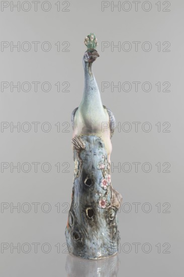 Peacock figurine, Chinese, Qing 1644–1911, 19th century; ceramics, colored porcelain; subject: peacock with peacock-feather ornament, plum trees, tree trunks, flowers; sculpture; height 54.5 cm, width 14.0 cm, depth 12.5 cm, weight 2.9 kg., A ceramic sculpture of a stylized bird perched on a textured trunk is shown against a plain gray background, the bird featuring a long neck and a small crest with eye-shaped details; the surface displays a mix of pale green, light blue, soft pink, muted purple, and touches of brown and beige, while the trunk shows darker blue-gray and brown tones with subtle floral decorations in pink and cream and areas of lighter green, and the sculpture has a glossy finish with visible hollows and carved details.