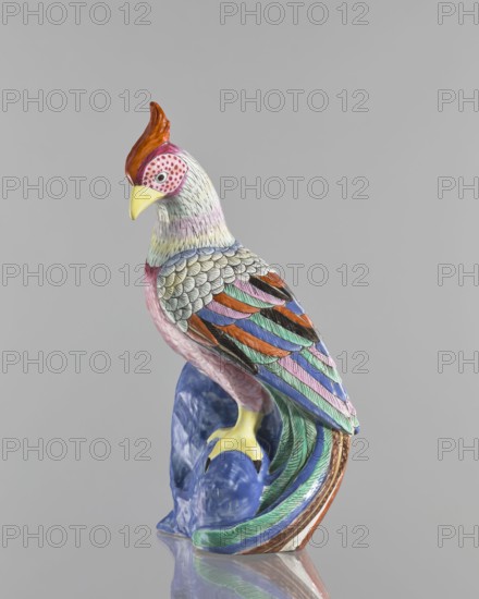 Rooster figurine, Chinese, Qing 1644–1911, 19th century, colored porcelain ceramic sculpture of a rooster with rocks; height 28.5 cm, width 14.5 cm; animal figurine bird/rooster iconography., A painted ceramic bird figurine perched on a glossy blue base, shown against a plain gray background; the bird has a red crest, a yellow beak, a pink face with dotted markings, and a pale neck with subtle pastel shading; its body and wings display a mix of colors including gray, pink, orange, green, blue, purple, black, and teal with detailed feather patterns; the legs and feet are yellow, and the base shows marbled blue tones with reflections on the surface beneath.