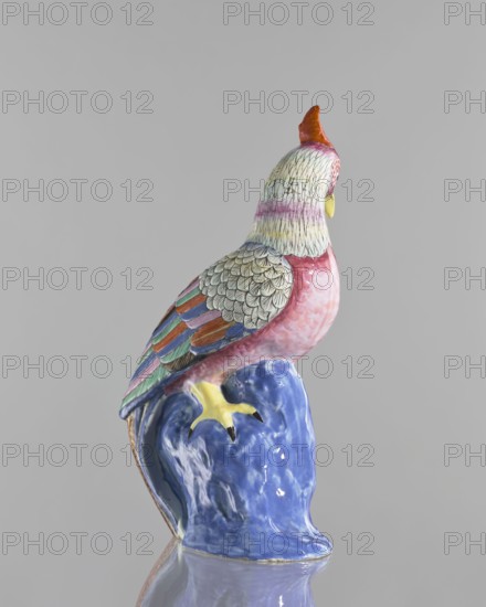 Rooster figurine, Chinese art, Chinese culture, Qing 1644–1911, colored porcelain, ceramics, animal figurines, birds, roosters iconography, rocks, 19th century, China, sculpture, height 28.5 cm, width 14.5 cm, A painted ceramic bird figurine perched on a glossy sculpted base is shown from a rear-side angle, with a background of light gray. The bird displays a palette including pink on the chest, cream and pale yellow on the head with fine dark line detailing, orange on the crest, and a yellow beak and claws. The wings show layered feather patterns in teal, green, blue, purple, orange, and tan with dark outlines, and the back feathers are cream with dark scalloped markings. The base beneath the bird is glazed in a deep blue with lighter highlights, and the surface beneath the object reflects the colors and shapes.