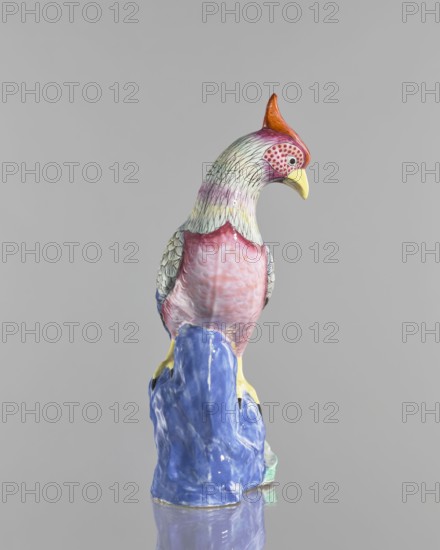 Rooster figurine, creator unknown, Chinese art, Chinese culture, Qing 1644–1911, colored porcelain, ceramics, animal figurine of a bird/rooster, depicted with rocks, 19th century, China, sculpture, height 28.5 cm, width 14.5 cm, A ceramic bird sculpture perched on a glossy rock-like base, shown against a plain light gray background. The bird has a crest colored orange and red, a yellow beak, a head and neck with pale green and lavender tones and fine dark feather lines, a face area with pink and red spotted pattern, wings with pale green and gray feather details, a breast shaded in pink and mauve, and yellow feet gripping a vibrant blue base that shows glossy reflections. The overall colors visible include orange, red, yellow, pale green, lavender, pink, mauve, gray, and blue.