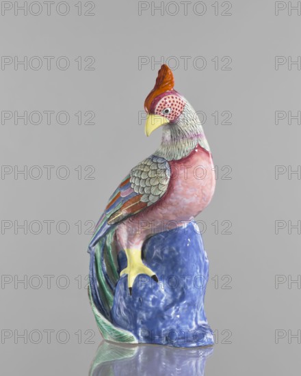 Rooster figurine, Chinese, Qing 1644–1911, 19th century; colored porcelain sculpture of a rooster with rocks; ceramics; animal figurine; birds; roosters iconography; height 28.5 cm, width 14.0 cm., A painted ceramic bird figurine sits on a glossy blue base against a plain light gray background, showing a bird with a red crest, yellow beak, and a face patterned with pink and white dots, a neck with fine dark lines on a pale background, a pink breast, multi-colored wing feathers in shades of green, blue, orange, and purple with scale-like detailing, yellow feet with dark tips, and a long tail with green and blue hues that curves beside the base; the base reflects on the surface beneath it.