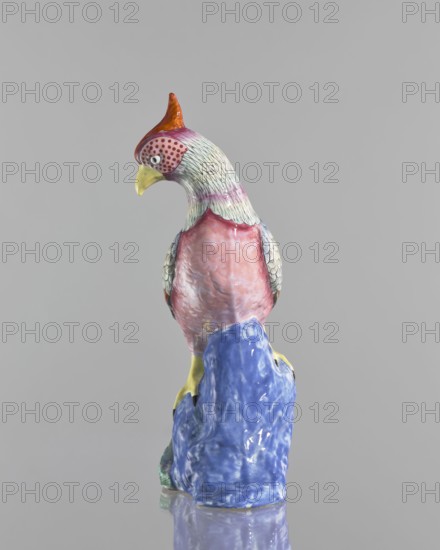 Rooster figurine, Chinese, Qing 1644–1911, 19th century. Colored porcelain sculpture of a rooster with rocks; height 28.5 cm, width 14.0 cm. Ceramic animal figurine., A ceramic bird sculpture perched on a sculpted base, shown against a plain gray background. The bird has a crest in orange and red, a yellow beak, and a head with pale greenish feathers and areas of red dotted patterning near the eye. The neck and upper chest show pale greenish feathers transitioning to pink and red tones on the body, with textured wing feathers in pale green and gray. The base beneath the bird is glossy blue with lighter and darker blue variations, and the bird's feet are yellow. The overall palette includes gray, orange, red, yellow, pale green, pink, and blue.