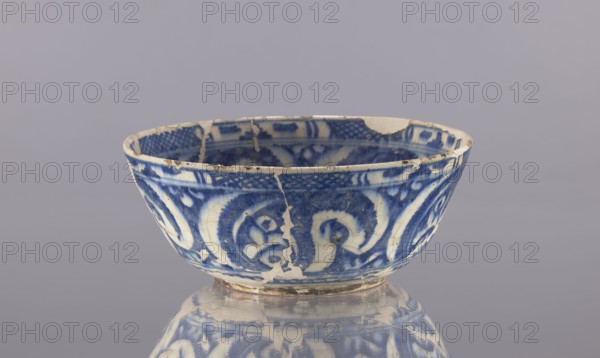 Bowl, Abdullazad Mirza Baba fl. ante 1939, revindication Kraków, Islamic art, Persian art, Persian culture, Qajar dynasty 1785–1925, glazed ceramics, utility ceramic bowl with plant motifs depicting everyday life in the Middle East, 19th century, Iran, vessel, height 9.5 cm, diameter 22.5 cm, A shallow ceramic bowl with a rounded shape sits against a plain light gray background, displaying a blue and off-white painted pattern on both its interior and exterior surfaces; the rim shows chips and small missing pieces, and there are visible cracks and areas of repaired or missing glaze. The blue decoration forms swirling floral and abstract motifs against the off-white base, and the bowl rests on a small foot ring that shows wear and discoloration; a reflective surface beneath the bowl produces a blurred mirror image that repeats the blue and off-white pattern.