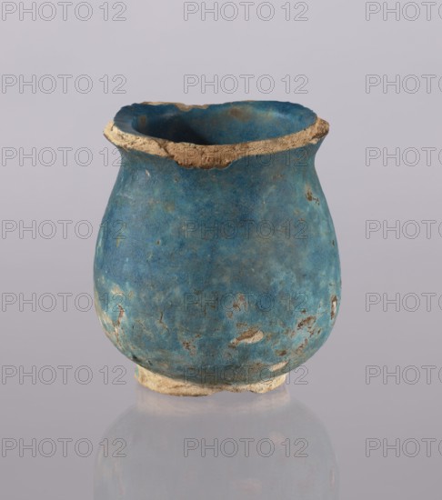 Jug, Ilkhanid period 1256–1335, Persian/Islamic ceramics, 13th–14th century Iran. Ceramic vessel cup/vase, used in everyday life in the Middle East; height 12.5 cm, diameter 11.5 cm, weight 0.378 kg., A small rounded vessel sits centered against a pale gray background, showing a worn blue surface with patches of lighter and darker blue and areas of brownish discoloration. The rim and base display exposed beige, sandy material where the blue surface is chipped away. The vessel has an uneven, slightly rough texture with some visible pitting and flaking, and the interior appears darker blue than parts of the exterior. The overall palette includes blue, beige, brown, and pale gray.