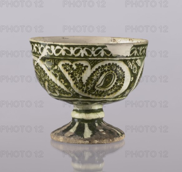 Cup, Abdullazad Mirza Baba fl. ante 1939, Islamic art, Persian art, Persian culture, Qajar dynasty 1785–1925, glazed ceramics, utility vessel with zoomorphic motifs, early 20th century, Iran; height 13.5 cm, diameter 11 cm, A ceramic pedestal bowl with a rounded body and flared rim displayed against a light gray background, decorated in a dark green and off-white palette with an intricate repeating leafy and paisley-like pattern around the exterior; the rim shows a few chips and cracks with exposed off-white clay, and the glaze on the surface has a slightly crazed texture. The stem and foot are painted with alternating dark green and off-white bands, and the foot shows worn, earthy discoloration at its edge; the bowl casts a soft reflection on the smooth surface below.