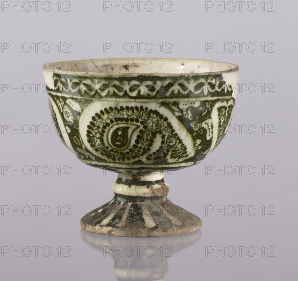 Cup, Abdullazad Mirza Baba fl. ante 1939, Islamic art, Persian art, Persian culture, Qajar dynasty 1785–1925, glazed ceramic vessel with zoomorphic motifs, utility ceramic, early 20th century, Iran; height 13.5 cm, diameter 11 cm., A small footed bowl with a rounded cup and flared pedestal, decorated with a repeating ornate pattern in dark green against a cream background, showing a few areas of glaze wear and darker spots; the rim is slightly chipped in places; the pedestal has vertical dark green stripes and a worn, earthy base with brownish discoloration; the surface reflects faintly on the pale lavender-gray surface beneath it.