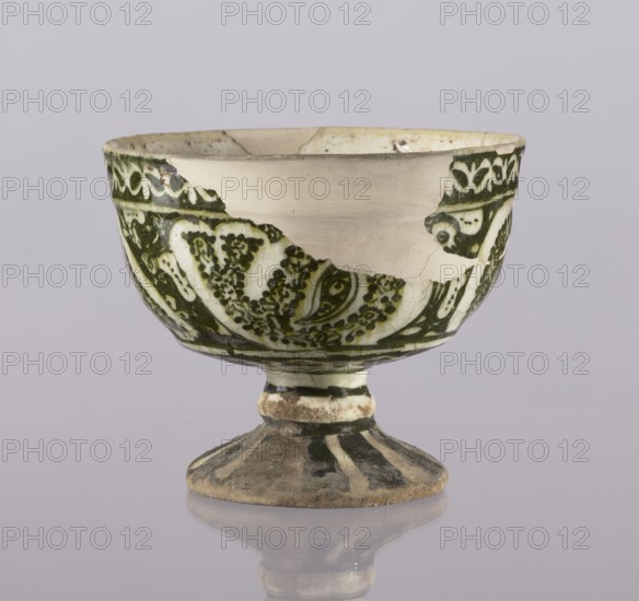 Cup, Abdullazad Mirza Baba fl. ante 1939, Islamic art, Persian art, Persian culture, Qajar dynasty 1785–1925, ceramics, glazed ceramics, utility ceramics, zoomorphic motifs, early 20th century, Iran, vessel, height 13.5 cm, diameter 11 cm, A ceramic footed bowl with a rounded cup and a flared pedestal base sits against a pale gray background, showing a glazed surface with areas of worn and chipped glaze. The bowl displays a decorative painted pattern in dark green and off-white on much of its exterior, with a large unpainted pale beige patch where glaze is missing; the green pattern includes stylized floral and leafy motifs and dotted textures. The rim and interior edge show aged discoloration in creamy beige and light brown, and the base and stem exhibit earthy brown and muted black areas with signs of surface wear and accumulated dirt. The piece casts a soft reflection on the glossy surface beneath it.