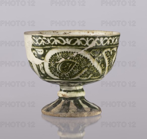 Cup, Abdullazad Mirza Baba fl. ante 1939, Islamic, Persian, Qajar dynasty 1785–1925, glazed ceramic vessel with zoomorphic motifs, utility ceramic, early 20th century, Iran; height 13.5 cm, diameter 11 cm., A footed ceramic bowl with a rounded cup and flared pedestal is shown against a pale grayish-lavender background, displaying a cream base color with dark green painted decorative patterns; the bowl surface has a band of interlaced vine-like motifs near the rim and a series of swirling floral and paisley-like designs around the body, with visible crack lines and a few repaired chips where the cream body shows through, and the pedestal foot has alternating dark green and cream vertical stripes and a slightly worn, earthy base that reflects faintly on the surface below.