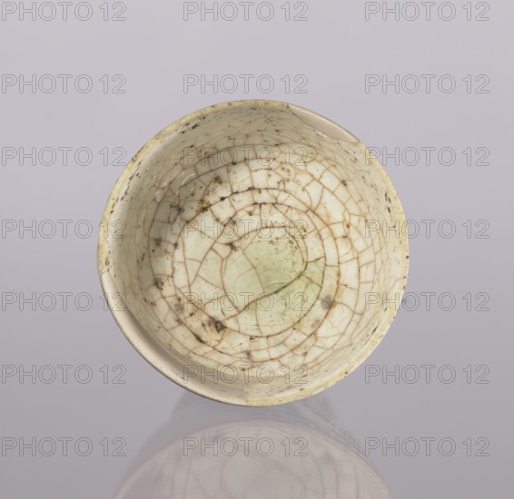 Cup, Abdullazad Mirza Baba fl. ante 1939, Islamic art, Persian art, Persian culture, Qajar dynasty 1785–1925, glazed ceramics, utility ceramic vessel with zoomorphic motifs, early 20th century, Iran, height 13.5 cm, diameter 11 cm, A shallow round bowl viewed from above with a crackled glazed surface showing an irregular network of fine lines and a few darker spots, primarily in shades of off-white, cream, beige, pale green, and brown, set against a soft pale grayish-lavender background that also reflects a faint mirrored image of the bowl.