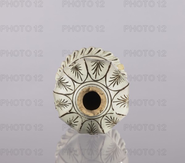 Bottle, Abdullazad Mirza Baba fl. ante 1939, Islamic art, Persian, Qajar dynasty 1785–1925, glazed ceramics, bottle fragment with diagonally knobbed foot edge. Decoration: bronze lines and stylized feathery twigs. 19th century, Iran. Vessel; height 19.0 cm, width 12.0 cm., A close-up of a round ceramic object viewed from one end, showing a circular central opening with visible rough interior edges, surrounded by a patterned surface with repeating fan- or leaf-like motifs in dark brown on a pale cream background; the rim around the opening has a golden-beige edge. The object has a scalloped outer edge with the same pale cream color and darker accents. The scene includes a reflective surface beneath the object that mirrors parts of the pattern. The background is a soft muted lavender-gray. Colors visible include cream, dark brown, golden-beige, and lavender-gray.
