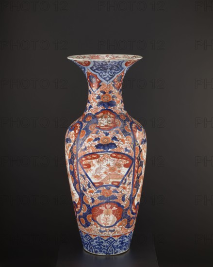 Vase, Japanese, Meiji period ca. 1870–1880, Arita. Colored porcelain Imari, decorated with plant ornaments and floral motifs—peonies, chrysanthemums, pine trees, momiji—and bird images including spotted pheasants and a hoo phoenix; komainu iconography and kachôga themes. Vessel, height 92.7 cm, diameter 37.5 cm, weight 22.5 kg., A tall decorative vase stands against a dark background, showing a glossy surface with intricate painted patterns in blue, red, white, and touches of gold or beige; the vase has a flared rim and a long neck leading to a wider body, with floral motifs, stylized leaves, and rounded medallion shapes arranged symmetrically around its form, alternating panels of dense blue ornamental designs and red floral scenes, and a band of blue patterned decoration near the base.