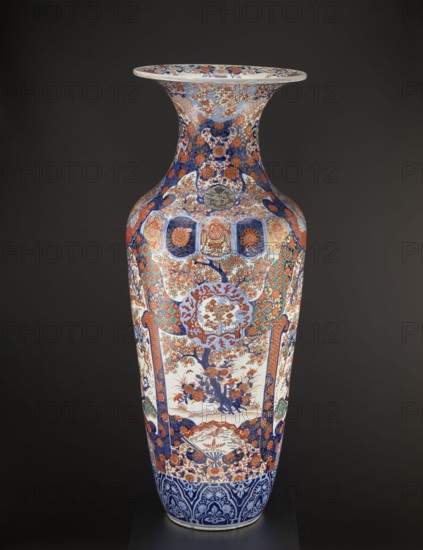 Vase, Fujisaki, Tahei,  craftsman, Japanese art, Japanese culture, Meiji 1868–1912, vases; subject: birds, floral decorations, plant ornaments, cherries, phoenixes iconography, turtles, chrysanthemums, gourds, fish, cranes, fans, dragons, butterflies; patterns: kikko, kojitsunagi, seigaiha; materials/technique: ceramics, colored porcelain, imari; sakura iconography, hoo phoenix. Ovoid vase with slender belly turning into a long neck with a flared spout. Four-reserve decoration with a cherry blossom tree and alternating phoenixes; multicolored background with geometric-floral patterns. ca. 1870–1880 Meiji Period, Japan; Hizen province, Arita. Vessel; height 122.0 cm, diameter 44.0 cm, weight 43.0 kg., A tall decorative vase with a flared rim stands against a dark background, covered in intricate painted patterns and motifs in a palette that includes blue, red, orange, white, green, and gold tones; the neck and shoulder display floral and circular medallion designs in blue and red with g...