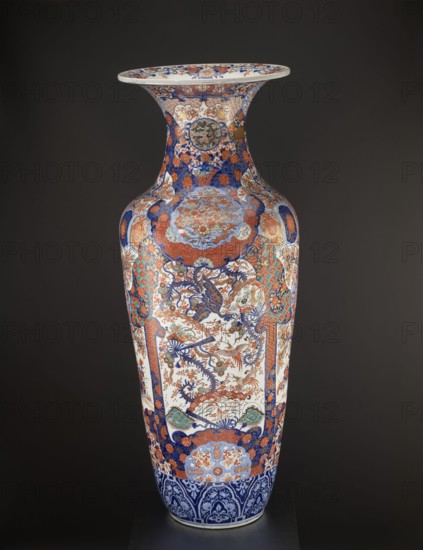 Vase. Maker: Fujisaki, Tahei,  craftsman. Provenance: unknown. Japanese art; culture: Japanese. Period: Meiji 1868–1912, ca. 1870–1880. Type: colored porcelain imari decorative vessel; ceramics; ovoid vase with slender belly turning into a long neck with a flared spout. Decoration: four-reserve decoration with a cherry blossom tree and alternating phoenixes; multicolored background with geometric-floral patterns; motifs include birds, floral decorations, plant ornaments, cherries, phoenixes hoo, sakura, turtles, chrysanthemums, gourds, fish, cranes, fans, dragons, butterflies; patterns kikko, kojitsunagi, seigaiha. Place of origin: Arita, Hizen, Japan. Function: vessel. Dimensions: height 122.0 cm; diameter 44.0 cm; weight 43.0 kg., A tall, slender ceramic vase with a flared rim sits against a dark background. The vase is richly decorated with intricate painted patterns and motifs in prominent colors of blue, orange, white, and touches of green and gold. The neck and shoulder feature d...