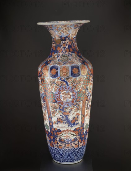 Vase, Fujisaki, Tahei,  craftsman, Japanese art, Japanese culture, Meiji 1868–1912, vases, birds, floral decorations, plant ornaments, cherries, phoenixes iconographic, ceramics, decorative vessels, colored porcelain, imari, sakura iconographic, turtles, chrysanthemums, gourds, fish, cranes, fans, dragons, butterflies, kikko pattern, kojitsunagi pattern, seigaiha pattern, hoo phoenix, Ovoid vase, slender belly turning into a long neck with a flared spout. Four-reserve decoration with a cherry blossom tree and alternating phoenixes; multicolored background with geometric-floral patterns. ca. 1870–1880 Meiji Period, Japan, Hizen province, Arita. Vessel; height 122.0 cm; diameter 44.0 cm; weight 43.0 kg., A tall decorative vase with a flared rim standing against a dark background, covered in intricate painted patterns and motifs in colors including blue, orange, red, white, green, and gold. The neck and shoulder area feature dense floral and geometric designs with alternating panels, whil...